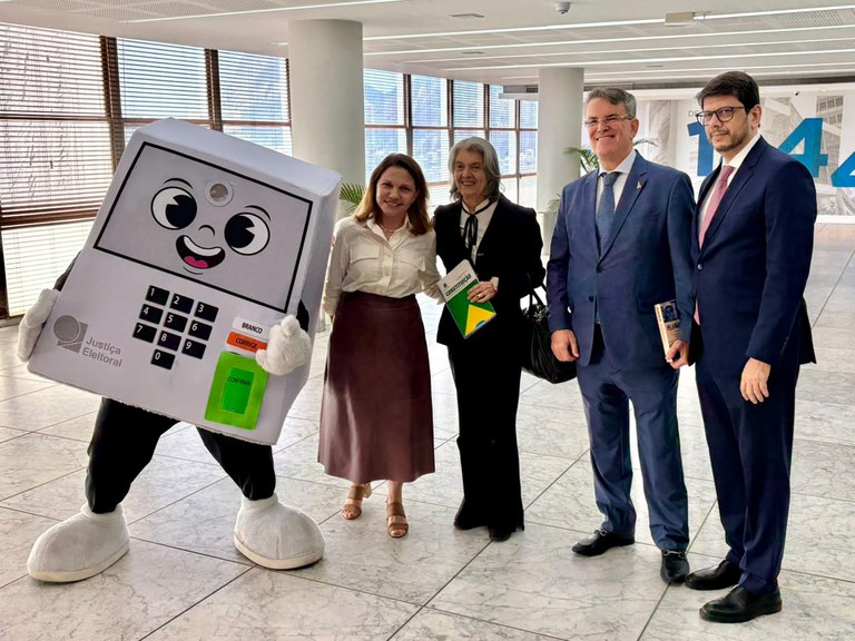 Fotografia num espaço interno com quatro figuras lado a lado. À esquerda, uma mascote em formato de urna eletrônica com o logótipo da Justiça Eleitoral. Ao lado,  Silvana Batini veste blusa clara e saia castanha, Cármen Lúcia usa terno preto segura a Constituição, e Cláudio Tavares e Flávio Paixão usam terno e gravata. Ao fundo, janelas de vidro e chão em mármore.