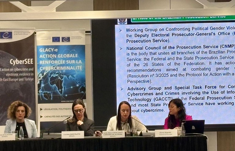 Quatro mulheres, sentadas lado a lado em uma mesa de trabalho, estão em um painel de evento. Elas olham para frente, enquanto a mulher na centro-direita fala ao microfone. No fundo, há um banner com o logotipo do Conselho da Europa e um telão exibindo tópicos em português sobre o Ministério Público Federal e combate à cibercriminalidade e violência de gênero. A imagem captura um momento de apresentação