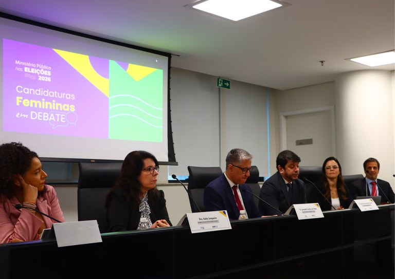 Seis palestrantes (3 homens e 3 mulheres) sentados em bancada com microfones. Ao fundo, tela exibe o texto: "Ministério Público nas Eleições 2026 - Candidaturas Femininas em Debate".