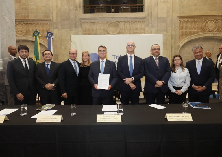 Sete homens e duas mulheres em trajes formais em pé atrás de mesa coberta com toalha preta e com copos e placas de identificação. Ao centro, o Presidente do TRE/RJ segura documento em papel em salão com duas bandeiras e painel institucional ao fundo