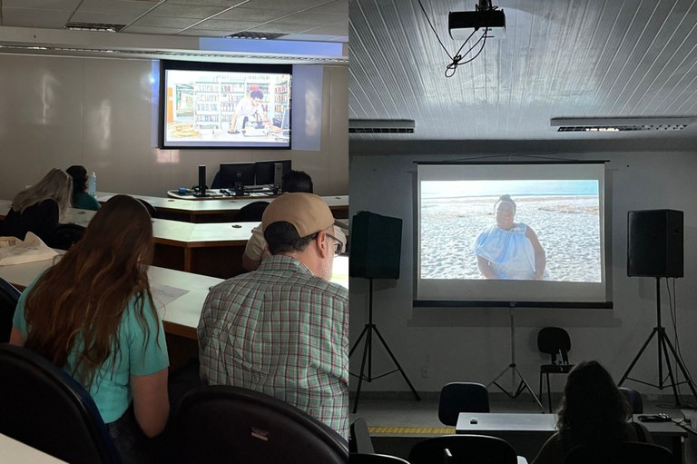 A imagem anexa é uma colagem lado a lado mostrando dois momentos distintos de projeção em sala de aula, provavelmente durante algum evento, aula ou seminário. Na imagem da esquerda, uma pequena plateia de estudantes (ou participantes), incluindo uma pessoa em primeiro plano com boné e camisa xadrez, assiste a um vídeo projetado numa tela menor, que parece mostrar uma pessoa em um ambiente com estantes de livros. A imagem da direita mostra uma projeção maior, numa tela suspensa com projetor no teto, onde é exibida a foto ou cena de uma pessoa sentada na areia de uma praia ou área costeira. Em ambas as cenas, a iluminação da sala é reduzida para facilitar a visualização da projeção, e a disposição sugere um ambiente universitário ou de treinamento.