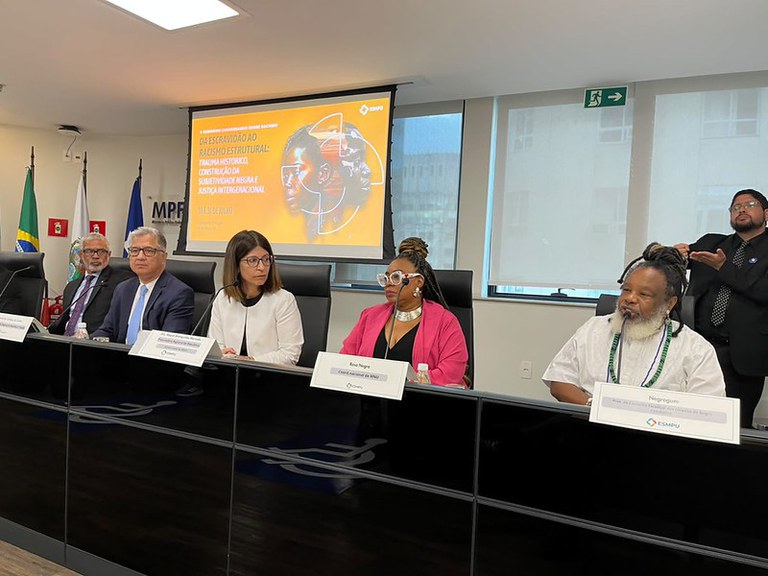 Fotografia de mesa de debate com cinco integrantes em bancada escura com microfones e placas de identificação. Ao fundo, tela projeta slide do "4º Seminário Conversando sobre Racismo", com o tema "Da Escravidão ao Racismo Estrutural: trauma histórico, construção da subjetividade negra e justiça intergeracional". À direita, intérprete de Libras em pé faz sinais. Na mesa estão presentes Rosa Negra, do MNU, e Negrogum, de túnica branca, entre outros. À esquerda, há bandeiras oficiais.