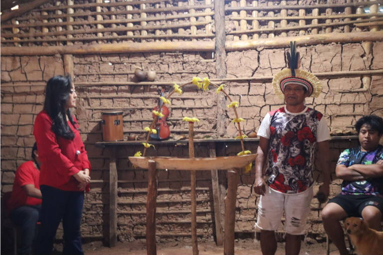 Foto mostra reunião no interior de uma das casas de reza na Terra Indígena do Jaraguá, um espaço de paredes de barro. Na foto, vemos um indígena com cocar em pé, ao lado de uma mulher oriental de camisa vermelha. Também vemos dois indígenas sentados, ouvindo o que é falado.