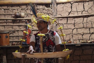 Homem indígena usando cocar posicionando-se atrás de uma estrutura ritualística de madeira decorada com penas amarelas, diante de uma parede de taipa.