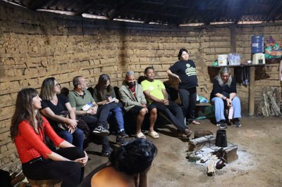 Pessoas sentadas em bancos ao longo de uma parede de barro dentro de uma estrutura tradicional, reunidas ao redor de uma pequena fogueira no chão.