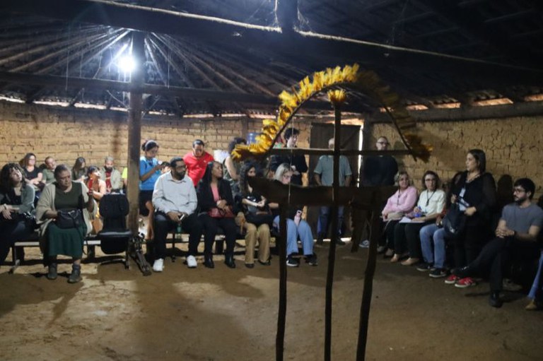 TIJaragua_3.jpg Grupo de pessoas reunidas em círculo dentro de uma casa de reza de taipa, observando um arco ritualístico ornamentado com penas amarelas em primeiro plano.