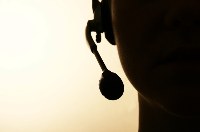 Fotografia em tons escuros mostra, em primeiro plano, o perfil de uma pessoa usando um headset com microfone. A imagem é capturada em contraluz, com o fundo iluminado em tom amarelado.