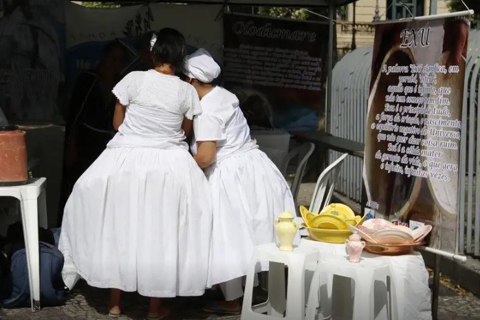 Fotografia de duas mulheres de costas para a câmera, vestidas em vestidos brancos, representando religiões de matriz africana.
