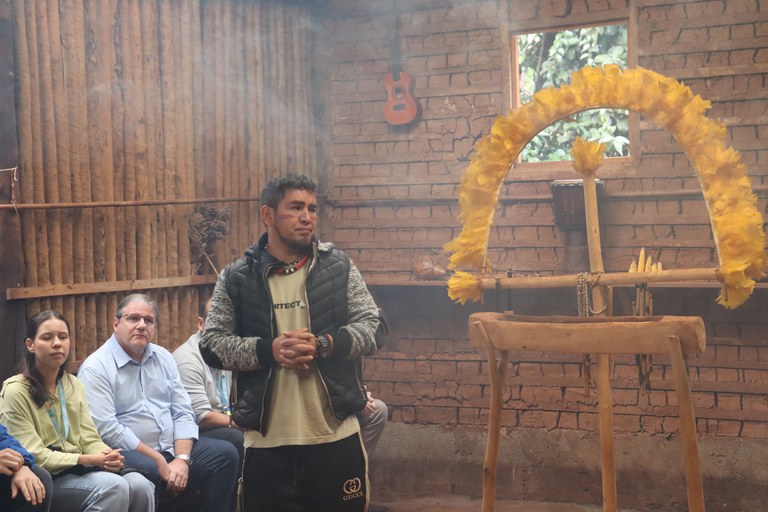 Foto mostra um homem indígena falando num local com paredes marrons, feitas de troncos e tijolos, com um grande cocar amarelo ao centro e um violão pregado numa das paredes.