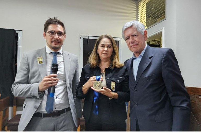 Foto mostra a procuradora Eugenia Augusta Gonzaga, uma mulher branca de roupa escura, com a medalha Manoel Fiel Filho nas mãos, no centro da foto, ao lado de dois homens de terno, um mais novo de óculos e terno cinza, e outro mais velho, de cabelos brancos e terno escuro.