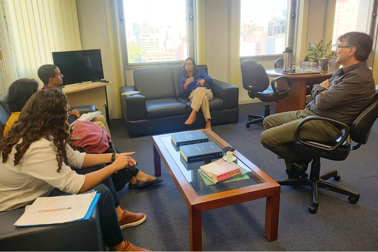 Sala com três estudantes sentados em um sofá, a procuradora Geisa de Assis Rodrigues sentada em outro sofá e o procurador Robério Nunes dos Anjos Filho sentado em uma cadeira de escritório de frente para os estudantes.