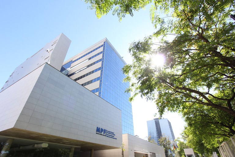 Sede da Procuradoria Regional da República da 4ª Região vista em um ângulo de baixo para cima. A construção tem duas torres, sendo que uma delas, onde ficam os gabinetes e a maioria dos setores administrativos, a fachada é espelhada. A imagem foi registrada em um dia com céu azul, e o sol aparece por entre as folhas de uma árvore que também está enquadrada na fotografia.