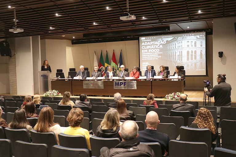 Mesa de debate em seminário do MPF sobre adaptação climática. Oito participantes em trajes formais sentam-se à bancada com microfones. Ao fundo, veem-se bandeiras e uma tela de projeção com o título Adaptação Climática em Foco. Parte do público também aparece de costas, sentado em poltronas.