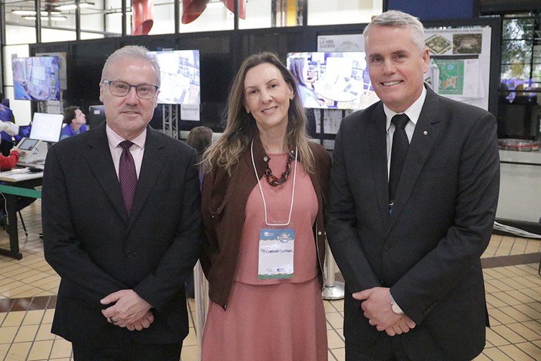 Foto da esquerda para direita de Claudio Fontella, Maria Emília Corrêa da Costa e Antônio Carlos Welter. Eles estão lado a lado e sorriem enquanto olham para a câmera. Os homens usam terno e gravata; a mulher, vestido rosa, casaco marrom e crachá no peito. Ao fundo, há grandes monitores que mostram o processo de auditoria das urnas.