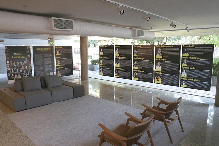 Imagem do foyer da PRR4, cujos vidros estão parcialmente cobertos pelos adesivos pretos com as artes da exposição. Aparece na imagem ainda um sofá cinza, duas cadeiras de madeira e um tapete cinza, centralizados na imagem e cercados pelos adesivos