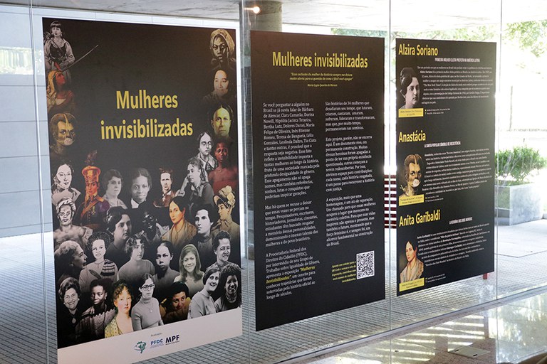 Foto horizontal de três banners com fundo preto e artes da exposição colados nos vidros do foyer da PRR4