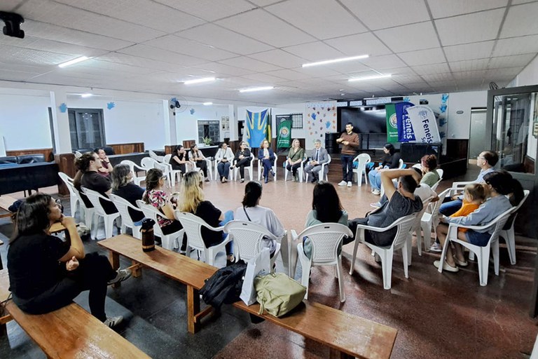 Fotografia em plano aberto em que cerca de 20 pessoas estão sentadas em cadeiras brancas de plástico arranjadas em um grande círculo. Ao centro, um homem em pé realiza gesticulações manuais (Libras). Ao fundo, há bandeiras e um painel. O ambiente tem iluminação clara, teto branco retangular e piso avermelhado. Em primeiro plano, observam-se bancos de madeira.