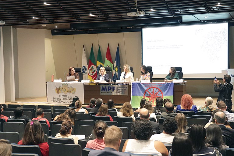Foto do auditório da PRR4, em que parte do público aparece de costas, sentado em poltronas. Ao fundo, no palco, aparecem uma mesa de autoridades de madeira composta por cinco mulheres e um telão.