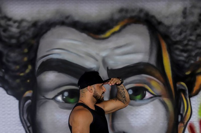 Foto mostra um homem em primeiro plano, de perfil, usando boné preto e camiseta sem mangas. Ele flexiona o braço esquerdo. Ao fundo, há um grande mural grafitado com o rosto de uma pessoa negra, olhos verdes e cabelo volumoso, que ocupa quase todo o fundo da cena. O contraste entre a pose do homem posicionado de lado e a expressão do rosto pintado posicionado de frente, cria um efeito visual de sobreposição de destaque.