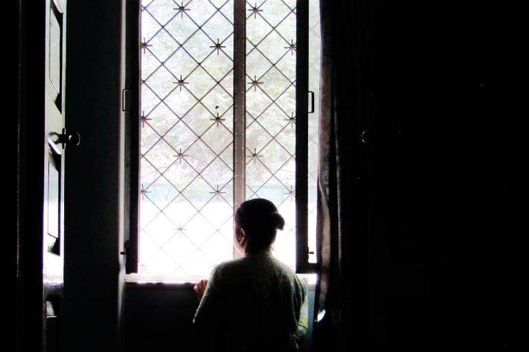 Foto mostra mulher em penumbra, de costas para o espectador e de frente para uma janela gradeada, por onde entra luz no cômodo