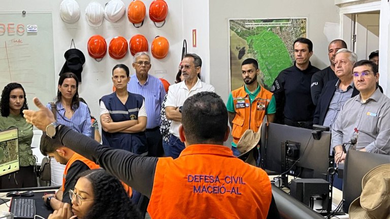 Técnicos da defesa civil, usando coletes laranjas, conversam com membros e servidores do MPF dentro da sala de monitoramento da Defesa Civil Municipal.