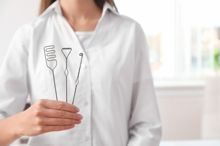 Close-up do tronco superior de uma profissional (possivelmente fonoaudióloga) vestindo um jaleco branco, segurando três instrumentos metálicos finos e especializados (provavelmente espátulas ou sondas) usados em terapia de fala ou avaliação orofacial.