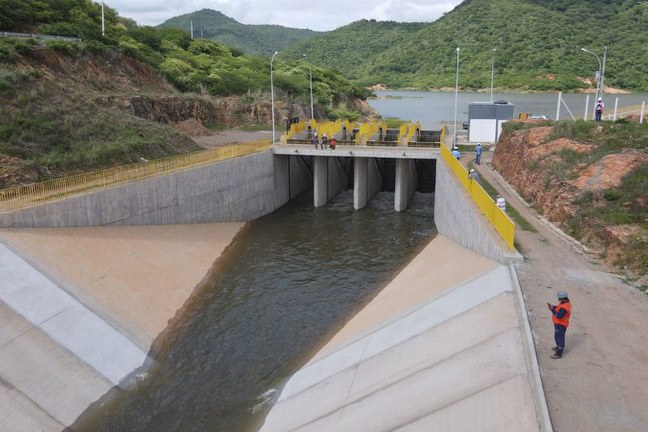 A imagem mostra uma barragem com comportas abertas, permitindo o escoamento da água. A estrutura é de concreto e está situada em uma área cercada por morros cobertos de vegetação. Algumas pessoas estão sobre a barragem e ao redor da área, aparentemente realizando inspeções ou trabalhos técnicos.