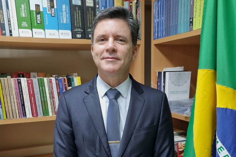 Foto do procurador regional da República Werton Costa, em plano médio curto. Ele aparece usando terno e gravata azuis e camisa branca, à frente de uma estante de livros, com uma bandeira do Brasil ao lado.