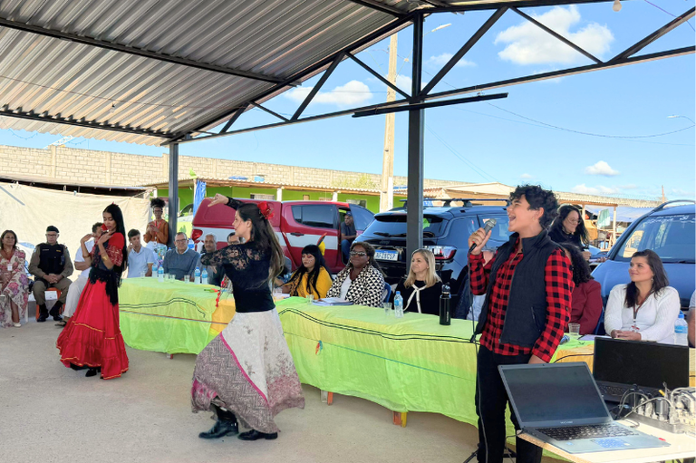 Foto mostra mais de uma dezena de pessoas sentadas à mesa de autoridades em ambiente aberto, mas coberto por telha de alumínio. Em frente à mesa, um homem canta com microfone na mão e duas mulheres, com saias longas, dançam.