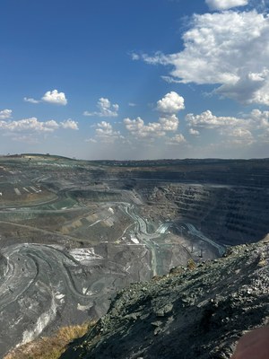 Fotografia em plano geral e perspectiva panorâmica de uma vasta mina de mineração a céu aberto sob um céu azul ensolarado com nuvens brancas dispersas. As paredes rochosas e escavadas da cratera formam degraus e caminhos sinuosos em tons de cinza, marrom e verde-azulado ao longo de toda a extensão do relevo. Em primeiro plano, no canto inferior direito, vê-se uma encosta íngreme de rochas e cascalho escuro, de onde se contempla a imensidão da cova de mineração ao fundo.