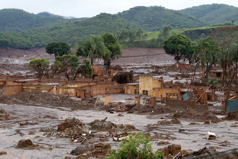 Fotografia tirada à distância mostra uma paisagem coberta de lama após o rompimento da barragem do Fundão, em Mariana (MG), com várias casas e construções destruídas.