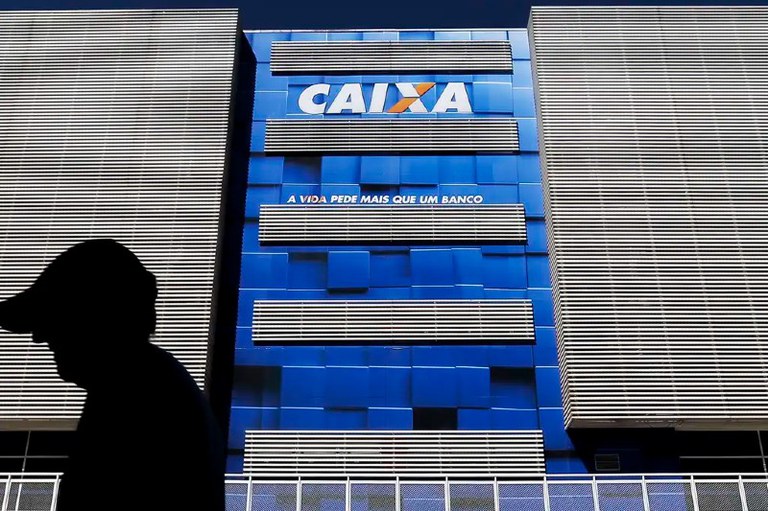 Foto mostra fachada de prédio da Caixa Econômica Federal. Um homem passa em frente ao edifício