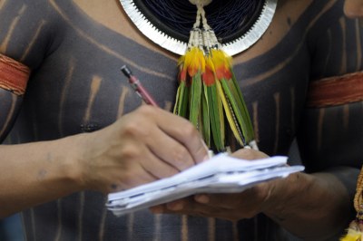 Fotografia mostra o torso de um indígena, com pintura corporal e adornos típicos, como colar de penas e braceletes. Ele está com um pequeno bloco ou caderno na mão, sobre o qual escreve com uma lapiseira marrom.