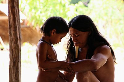 Homem indígena adulto agachado em frente a criança indígena, ambos aparentemente nus e a criança com colar. O adulto tem cabelos longos e usa o ’m’berpót’, ornamento labial longo feito de madeira introduzido no lábio inferior.