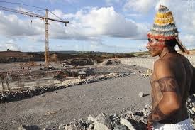 Foto de indígena da etnia Munduruku, com pinturas corporais e o cocar de penas típico desse povo, que observa o canteiro de obras da usina de Belo Monte durante manifestação em maio de 2013.