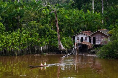 Foto de duas pessoas em uma canoa em rio, próximas a outras três pessoas que estão em frente a um conjunto de duas casas de palafita na beira do rio. O rio e as casas são cercados de floresta densa, de uma cor verde bem vívida.