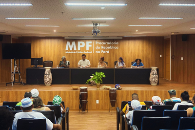Foto de seminário no auditório do MPF em Belém, que é todo em madeira clara e tem a logo da instituição ao fundo. Na foto, há uma mesa de palestrantes com cinco pessoas,  uma delas falando ao microfone.