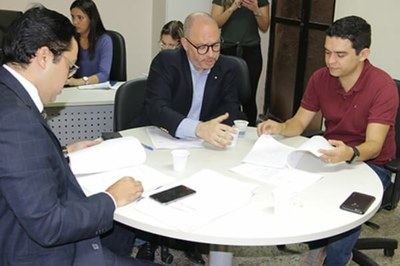 Três homens sentados a uma mesa lêem documentos. Ao fundo, três mulheres usando computador e celulares.
