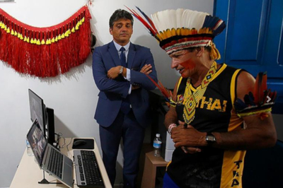 Homem indígena com cocar de penas coloridas na cabeça está em frente a um notebook, enquanto é observado por um homem vestindo paletó e gravata