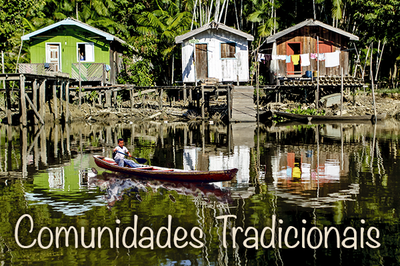 Fotografia de casas na beira de um rio, com uma pessoa sobre uma canoa, em primeiro plano, além de vegetação ao fundo. Na parte inferior da imagem tem o texto "Comunidades Tradicionais".