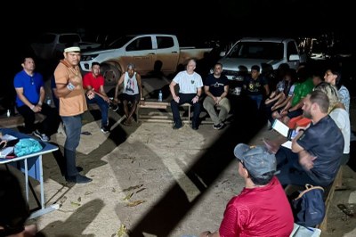 Homens e mulheres, entre eles indígenas, reunidos em área aberta, à noite, com chão de terra batida, sentados sobre bancos de madeira, tendo um indígena em pé, a falar com os demais presentes