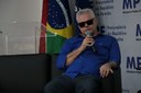 José Antônio Freire, presidente do Instituto dos Cegos da Paraíba. José está sentado em um sofá, segurando o microfone enquanto realiza sua fala. Ele usa óculos escuros e veste uma camisa azul com uma calça cinza. Em segundo plano, aparecem as bandeiras da Paraíba, Brasil e MPF.