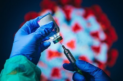#pracegover: foto retangular das mãos de um profissional de saúde segurando um frasco de vacina e uma seringa, com a representação de um coronavírus ao fundo