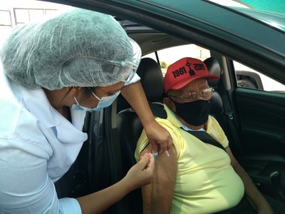 A imagem é uma foto de um senhor idoso que está sentado dentro de um carro sendo vacinado por uma enfermeira contra a covid-19.