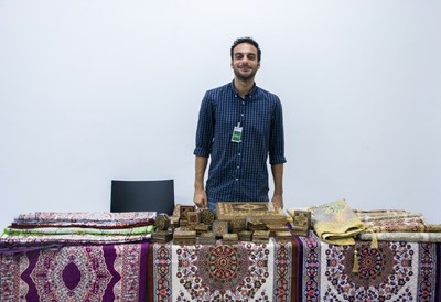 A imagem mostra um homem em pé, atrás de uma mesa, sobre a qual estão espalhados tapetes coloridos.