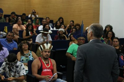 A imagem mostra pessoas olhando atentamente para um homem de paletó que está falando para a plateia.