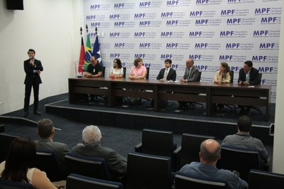 A imagem mostra o auditório do MPF onde vemos uma mesa composta pelos representantes dos órgãos participantes da reunião. Em pé, segurando um microfone, à esquerda, está o procurador Guilherme Ferraz falando para o público presente.  