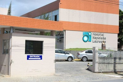A foto mostra a fachada do Hospital com a guarita, onde se lê a palavra 'ENTRADA" e um portão de correr na cor branca. Aparecem dois carros estacionados no jardim do hospital. Na parede da frente do edifício aparece o nome Hospital Napoleão Laureano.