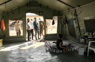 Foto do interior de um abrigo usado por migrantes tendo uma mulher sentada num colchão no chão, mesa e cadeira, e algumas pessoas na porta olhando para dentro do abrigo. 