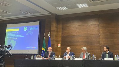 #pracegover: banner com foto do evento tendo à direita um telão reproduzindo o banner do evento, e à esquerda, a mesa com o PFDC Carlos Vilhena e outros participantes.